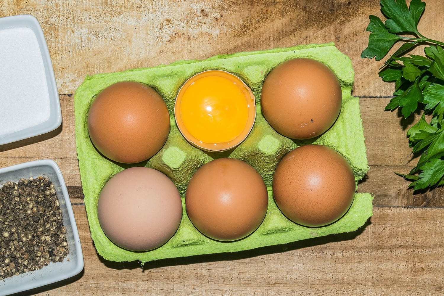 Enam butir telur ayam cokelat dalam wadah karton hijau, satu di antaranya terbelah menampakkan kuning telur